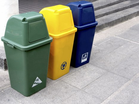 The image shows three upright wheelie bins arranged side by side on a paved sidewalk area outside a building, likely near a waste collection point in Cranford. The first bin on the left is green, featuring a simple, smooth plastic surface with a slightly rounded lid and a small triangular recycling or waste management symbol on the front. The middle bin is yellow, with a similar plastic structure and a slightly more prominent lid, and it displays a black recycling symbol consisting of three arrows forming a triangle on its front face. The bin on the right is dark blue, with a matte finish, a matching lid, and a white recycling icon printed on the front. All three bins are positioned in front of a grey, textured paving surface, with a slight shadow indicating daylight. The background includes a portion of a dark, possibly corrugated, exterior wall or pavement edge, suggesting an urban or commercial setting. These bins are representative of waste or rubbish collection containers used by offices or businesses, suitable for general disposal or recycling purposes in Cranford or nearby areas, and are connected to professional rubbish removal services such as those offered by Office Clearance Cranford.