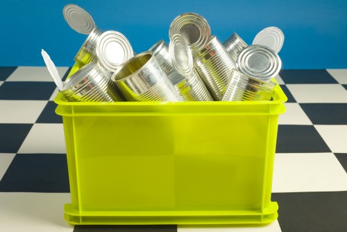 A bright yellow plastic bin filled with empty, silver metallic tin cans with smooth, cylindrical surfaces and open tops, some cans showing inward bent rims. The cans are stacked loosely inside the bin, which is positioned on a checkered black-and-white floor. The background features a solid blue wall, suggesting an indoor setting such as a recycling area or waste collection point. The image reflects waste management and rubbish removal services provided by Office Clearance Cranford, focusing on the collection and disposal of metal containers. The lighting is even, highlighting the reflective surfaces of the cans and the vibrant yellow of the bin, with no other objects or environmental details visible in this clean, straightforward scene.