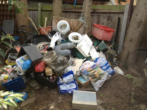A pile of mixed household waste and rubbish situated outdoors on a patch of bare soil, adjacent to a wooden fence and a couple of wooden posts. The collection includes various discarded items such as a black plastic storage box, several white and grey plastic pipes and components, a red plastic storage container, and crumpled paper and plastic bags. There are also cardboard boxes, some filled with miscellaneous debris, and an assortment of plastic packaging and wrapping material. Items are haphazardly stacked and scattered, with some leaning against the fence or partially covering each other. The overall scene appears to be part of an outdoor rubbish clearance, typical of household waste accumulation awaiting removal. The natural lighting suggests daytime, with some shaded areas cast by nearby trees and fencing, characteristic of a residential garden or backyard in Cranford, London.