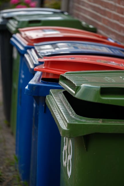 A row of large, rectangular wheelie bins lined up outdoors along a pavement, positioned side by side in front of a brick wall. The bins are made of sturdy plastic with textured, ribbed lids, each pain