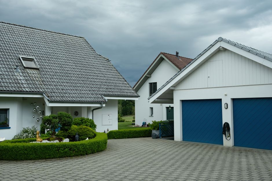 The image depicts the interior of a garage with open roller shutter doors revealing an outdoor area. Inside the garage, the concrete floor is partially covered with scattered debris, including small b