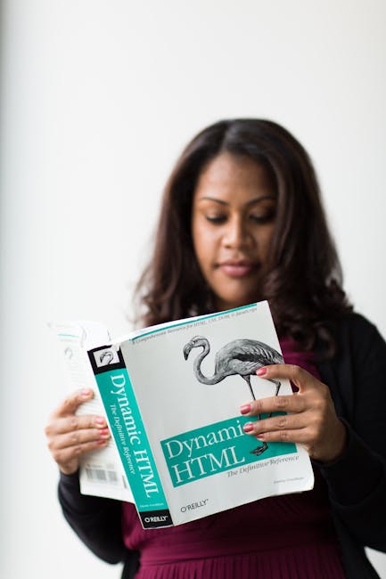 A woman with dark, wavy hair and medium skin tone is seen reading a book titled 'Dynamic HTML' against a plain white background. She is wearing a black blazer over a burgundy top. Her facial expressio
