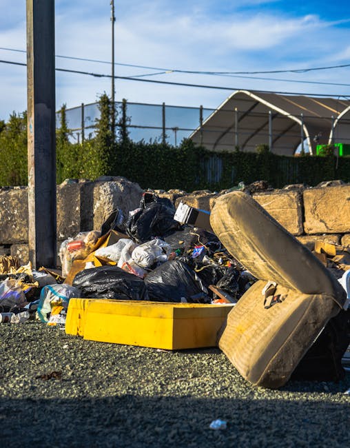 A narrow back alleyway between two brick and corrugated metal walls, both covered in graffiti, with scattered rubbish and discarded household items along the uneven, muddy ground. Visible objects incl