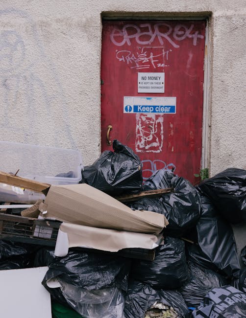 A pile of black rubbish bags and discarded packaging materials stacked against a weathered red metal door with graffiti and various stickers, including a white sign reading 'No Money' and a blue and w