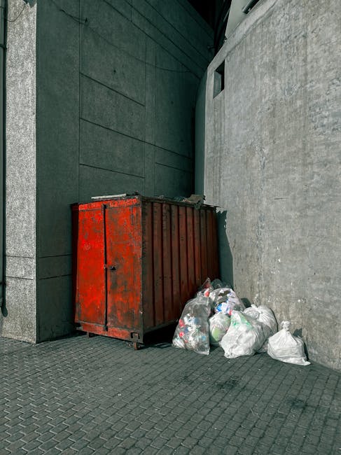 A pile of black rubbish bags and discarded packaging materials stacked against a weathered red metal door with graffiti and various stickers, including a white sign reading 'No Money' and a blue and w