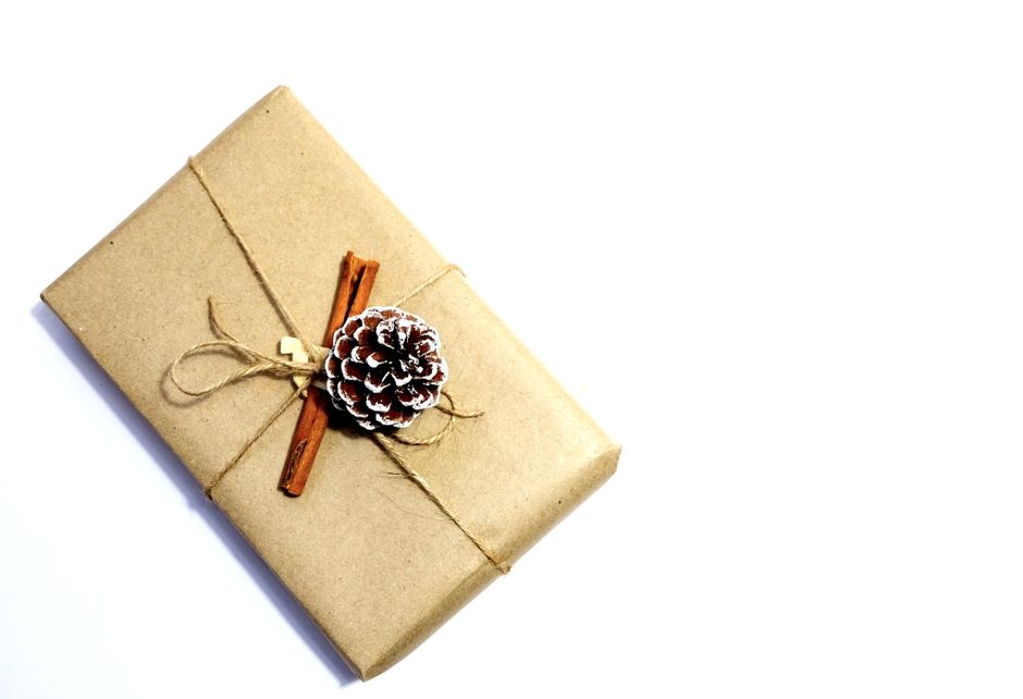Three small gift boxes decorated for Christmas are arranged on a plain white surface. The top box is wrapped in brown paper, adorned with a beige ribbon and a white wooden snowflake ornament, with spr